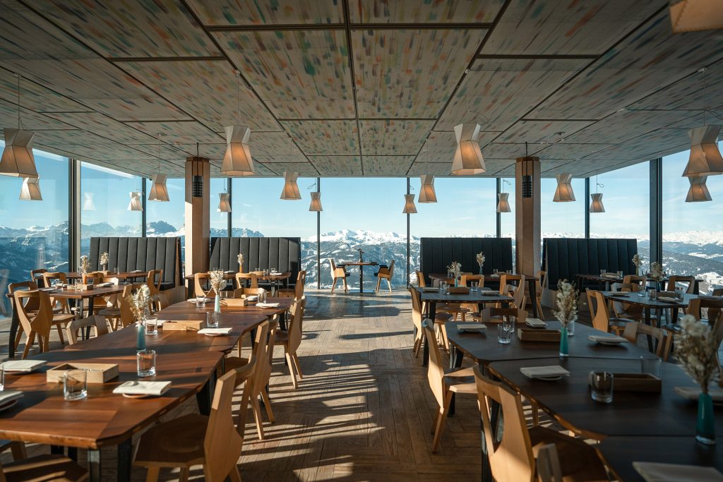 Modern alpine restaurant with panoramic mountain views, wooden tables, and stylish pendant lighting.